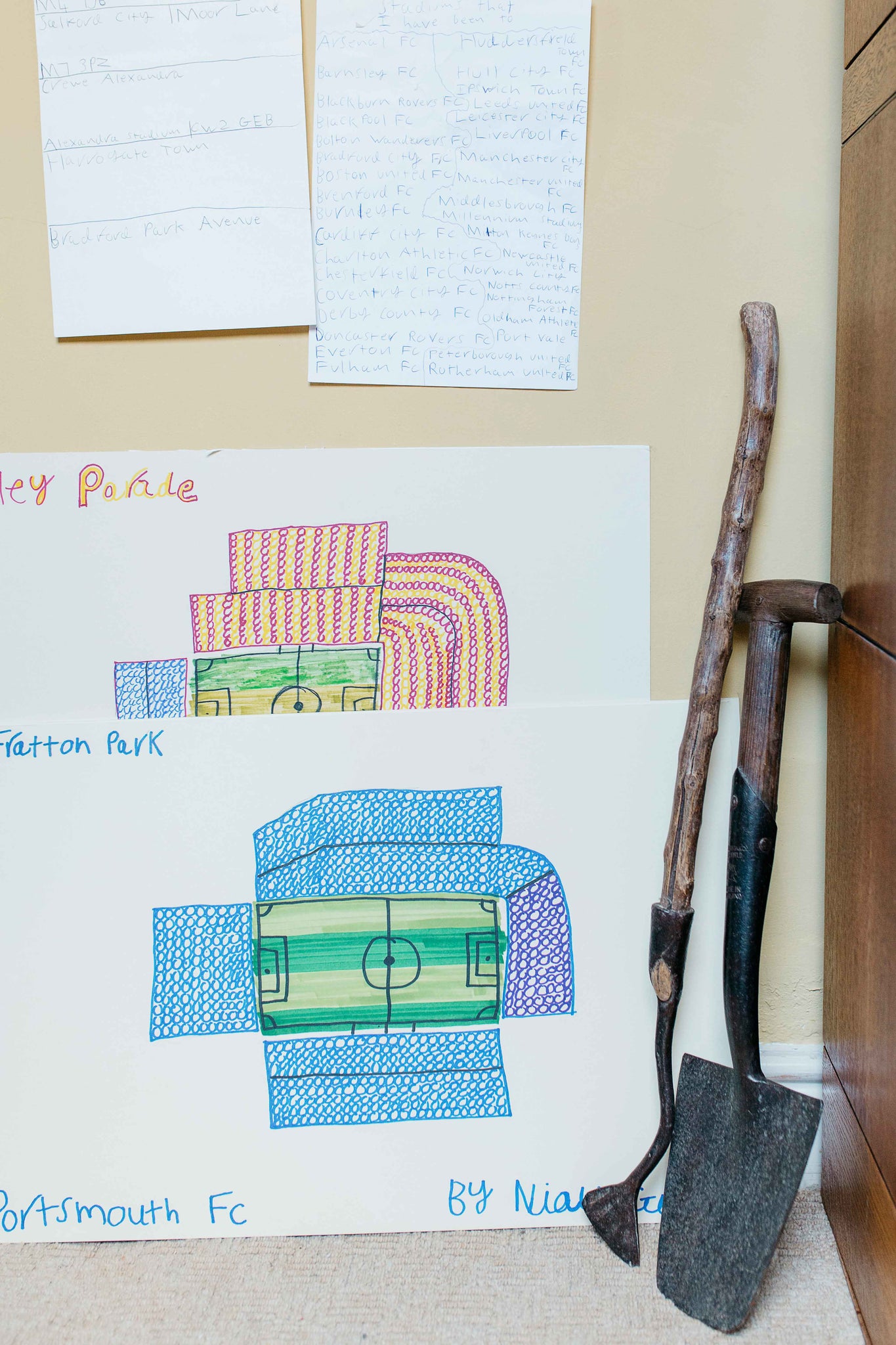 Two hand drawn football stadium drawings sit next to two gardening tools against a wall